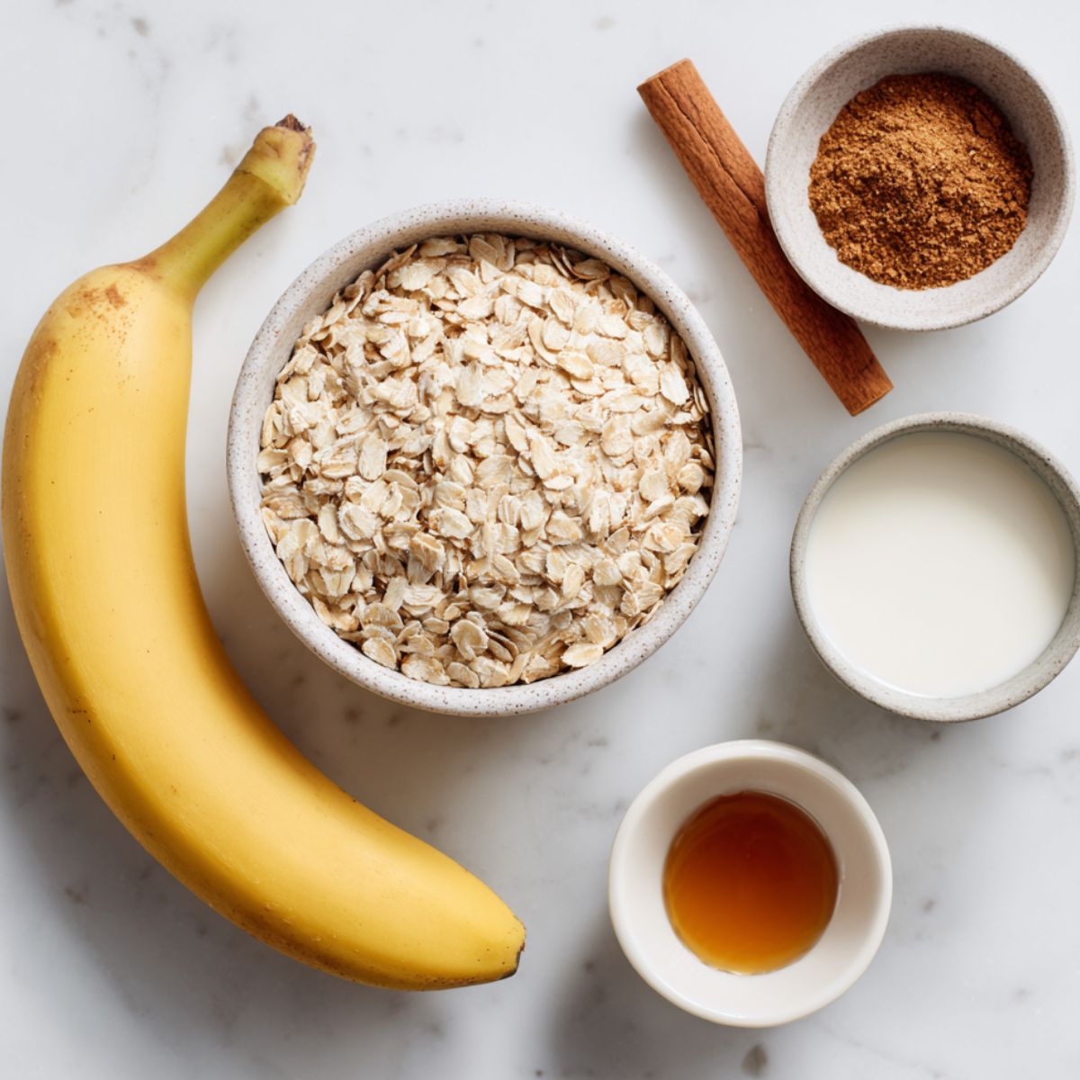 Rolled oats, banana, cinnamon, maple syrup, and milk arranged casually on a white kitchen counter for cinnamon roll baked oatmeal.