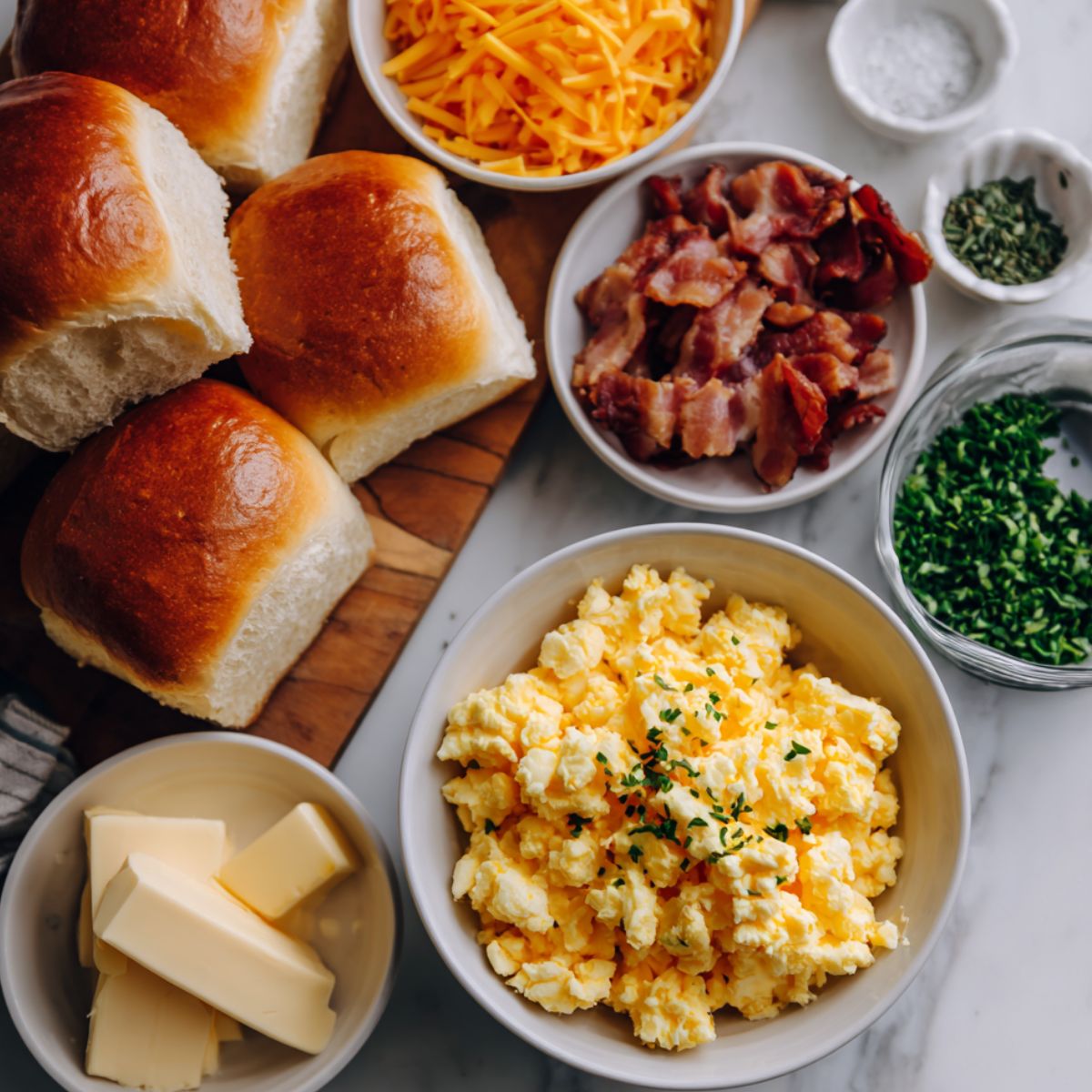 Flat lay of Hawaiian rolls, scrambled eggs, bacon, cheese and butter on a white kitchen counter.
