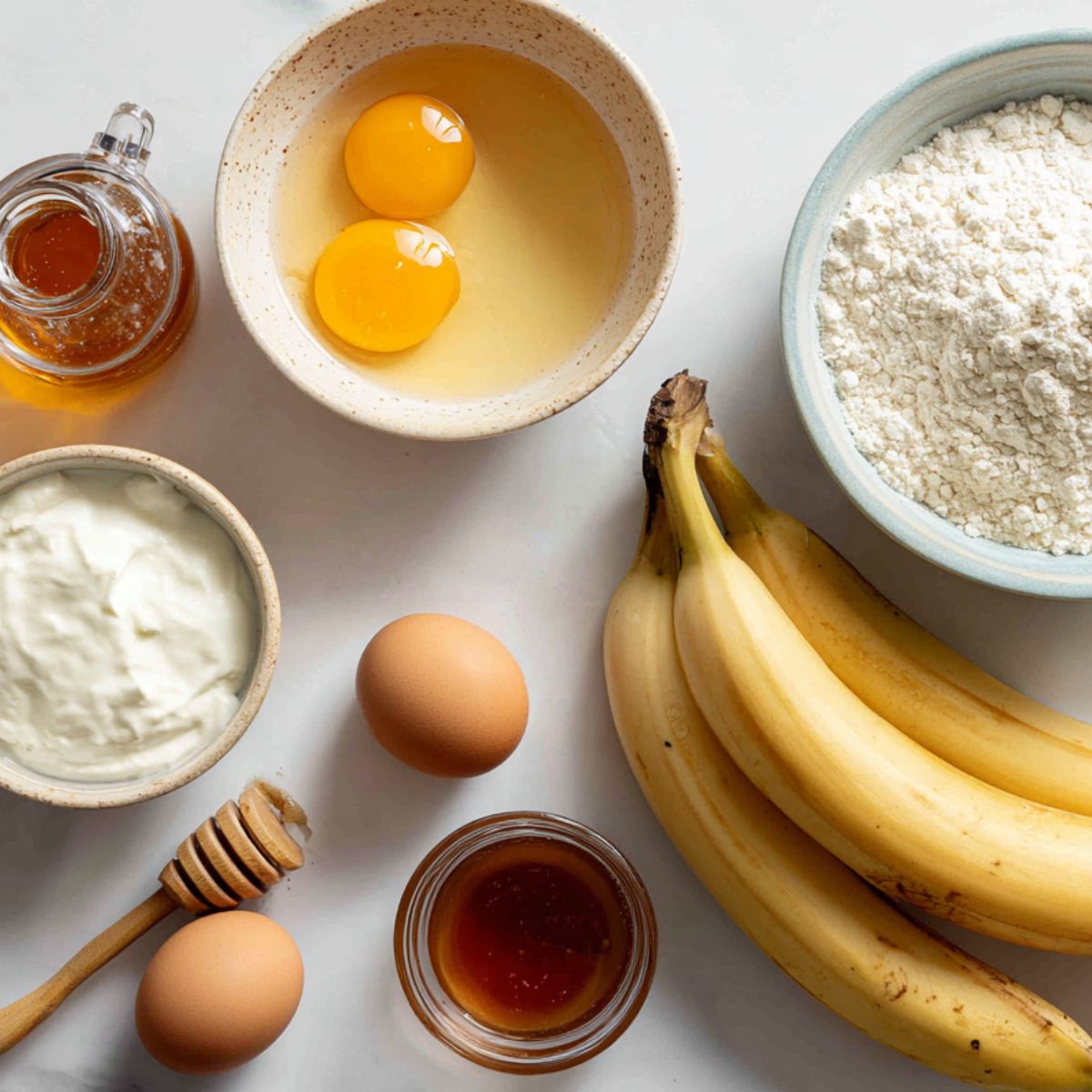 Flat lay of ripe bananas, Greek yogurt, eggs, honey, and flour on a white kitchen counter.
