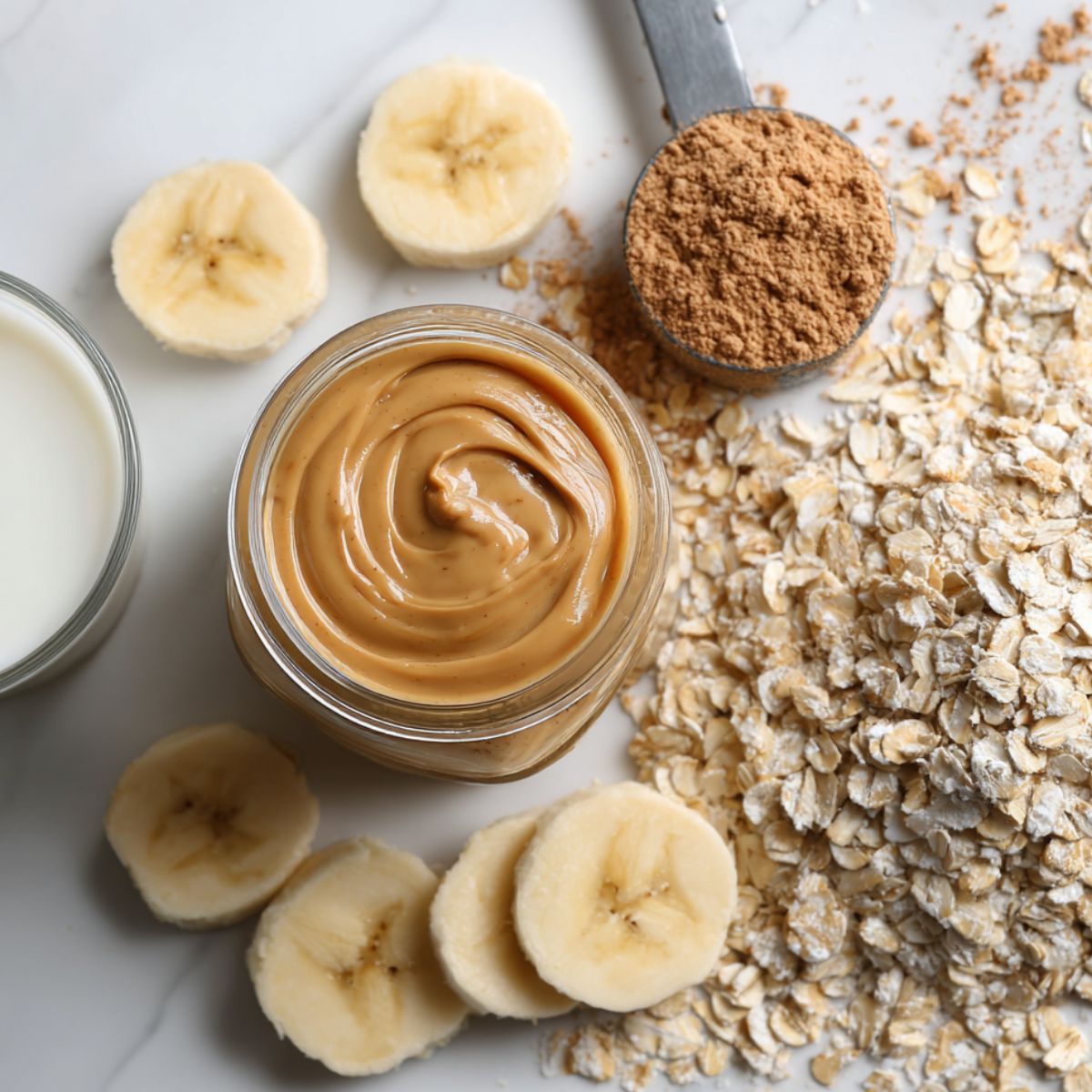 Homemade smoothie ingredients including banana, peanut butter, oats, protein powder, and milk on a white kitchen counter, top view.