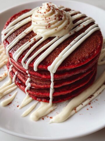 Overhead photo of a homemade stack of red velvet pancakes drizzled with cream cheese glaze on a white kitchen counter.