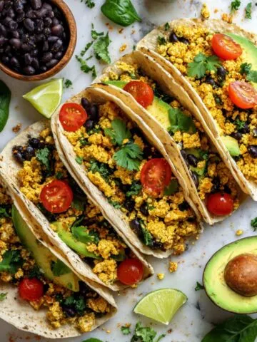 Overhead shot of four vegan breakfast tacos filled with tofu scramble, black beans, avocado, tomatoes, and cilantro on a white kitchen counter, with toppings scattered naturally