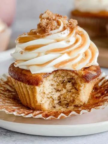 Homemade cinnamon roll cupcakes on a white counter, with imperfect frosting and a half-eaten one
