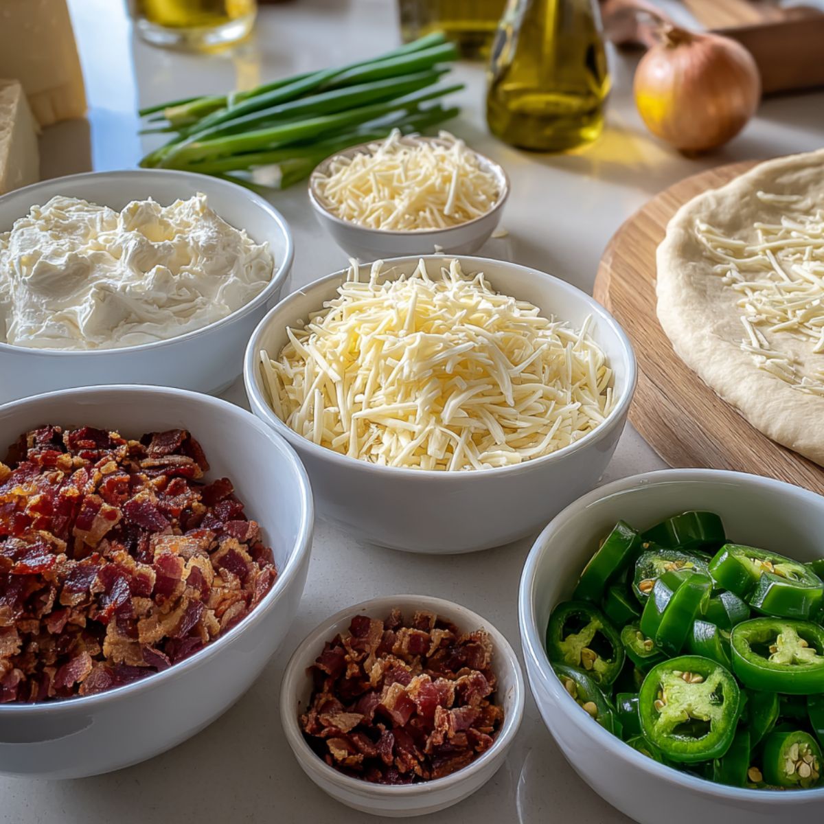 Overhead photo of homemade jalapeño popper pizza ingredients on a white kitchen counter including cheeses, jalapeños, bacon, green onions, and pizza dough.