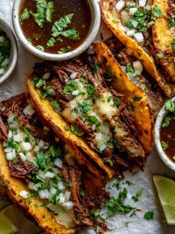 Homemade slow cooker birria tacos with consomé and toppings on a white kitchen counter