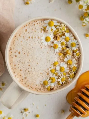 Homemade chamomile tea latte in a rustic mug with chamomile flowers and honey on a white kitchen counter, photographed from above.
