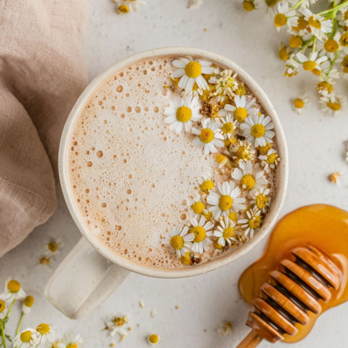 Homemade chamomile tea latte in a rustic mug with chamomile flowers and honey on a white kitchen counter, photographed from above.
