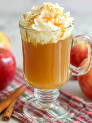 op-down view of a mug of homemade caramel apple cider with whipped cream, caramel drizzle, and a cinnamon stick on a white kitchen counter.