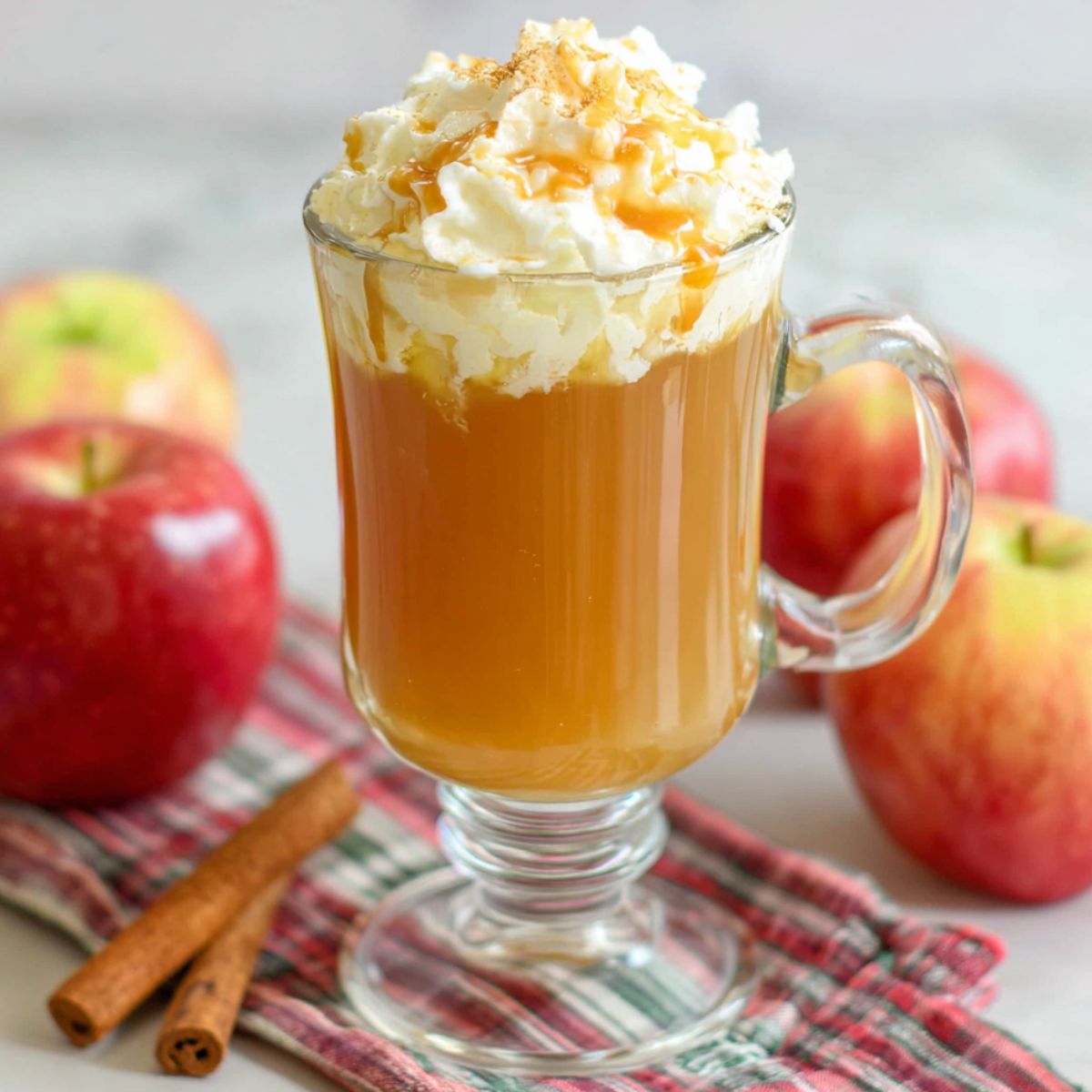 op-down view of a mug of homemade caramel apple cider with whipped cream, caramel drizzle, and a cinnamon stick on a white kitchen counter.