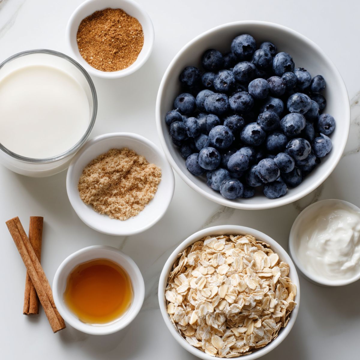 Top-down view of rolled oats, blueberries, milk, and yogurt laid out casually on a white kitchen counter for making Blueberry Pie Overnight Oats.