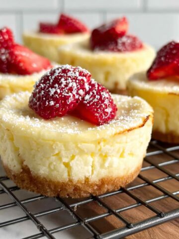 Flatlay of cheesecake cupcake ingredients on a white kitchen counter, showing cream cheese, eggs, butter, and graham cracker crumbs.