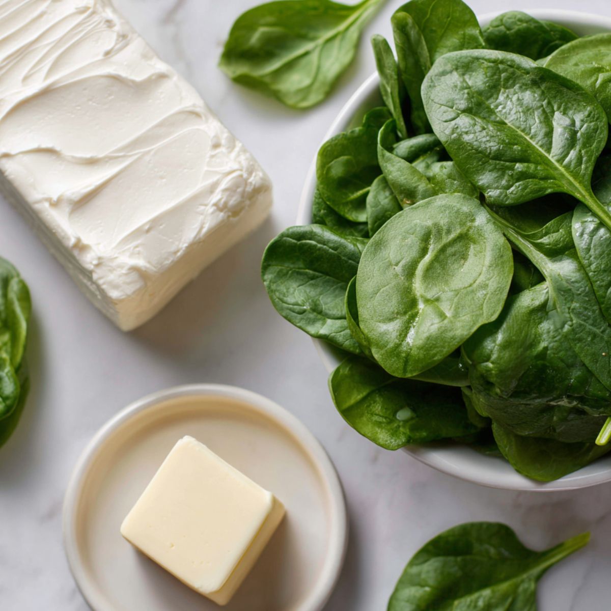 Fresh spinach, butter, and cream cheese arranged casually on a white kitchen counter, photographed from above