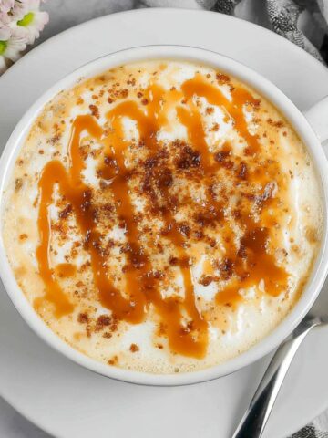 Homemade caramel brulée latte in a ceramic mug on a white kitchen counter, topped with whipped cream and caramel drizzle.