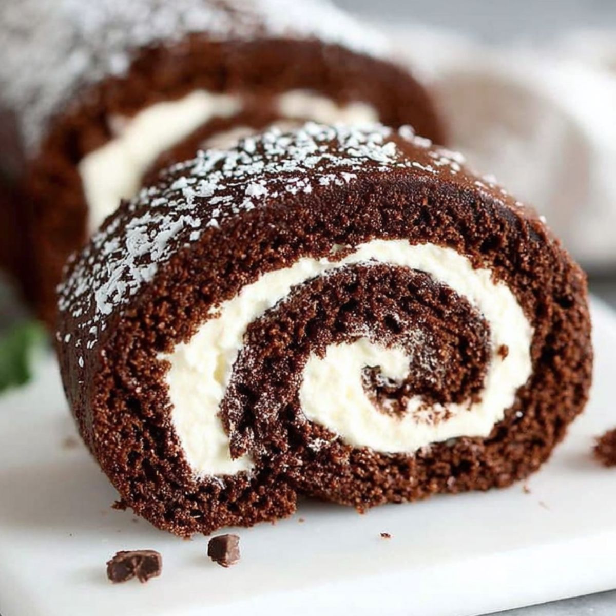 Homemade chocolate swiss roll cake with cocoa sponge and cream filling on a white kitchen counter, dusted with powdered sugar.