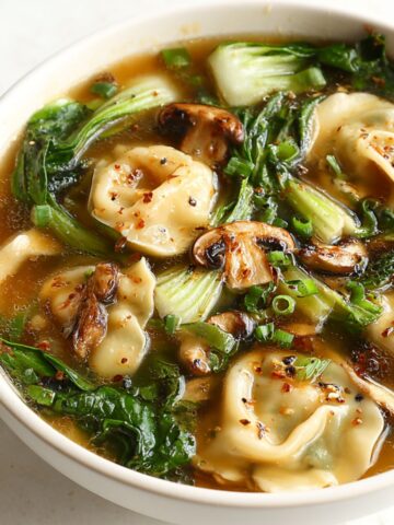 A warm bowl of potsticker soup with dumplings, bok choy, and mushrooms on a white kitchen counter.