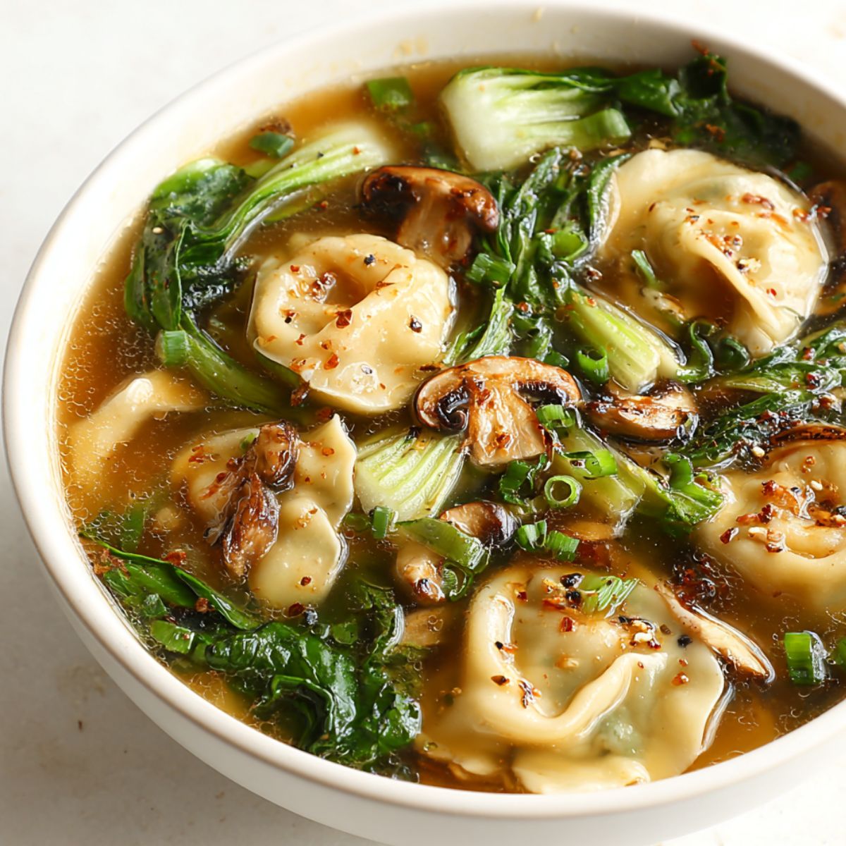 A warm bowl of potsticker soup with dumplings, bok choy, and mushrooms on a white kitchen counter.