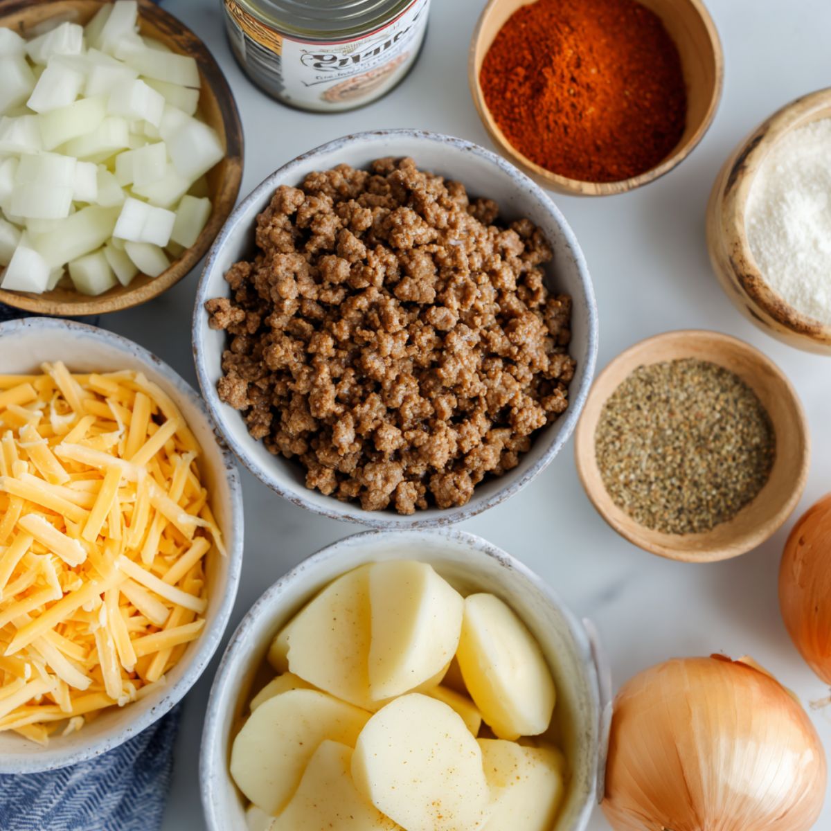 Flat lay of raw ingredients for hobo casserole including ground beef, sliced potatoes, onion, cheddar cheese, soup can, and spices on a white kitchen counter.