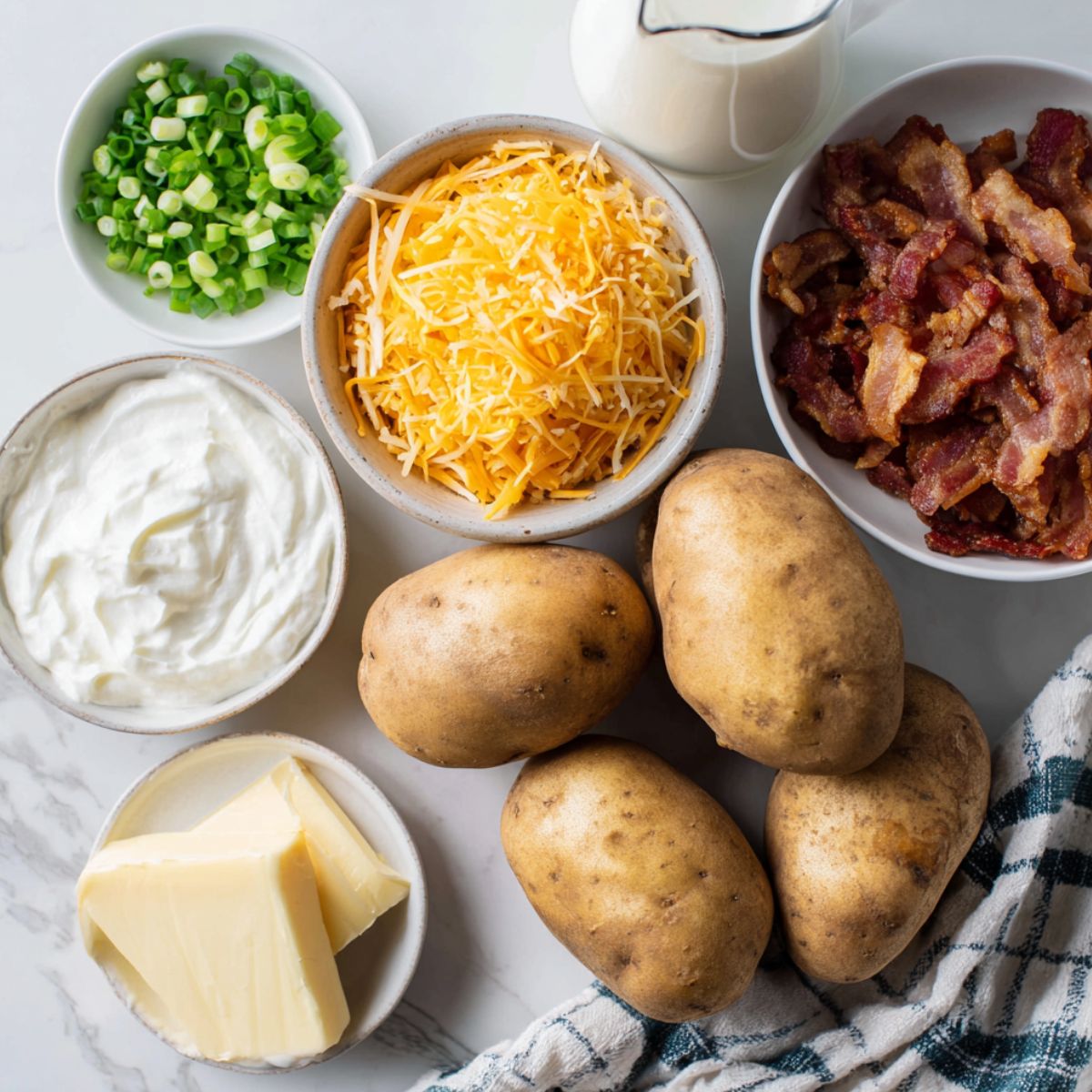 Overhead shot of ingredients for homemade loaded mashed potato casserole on a white counter — potatoes, cheese, bacon, sour cream, and green onions